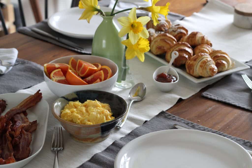 Easter brunch table with bacon, eggs, oranges, daffodils in a vase, and croissants with jam