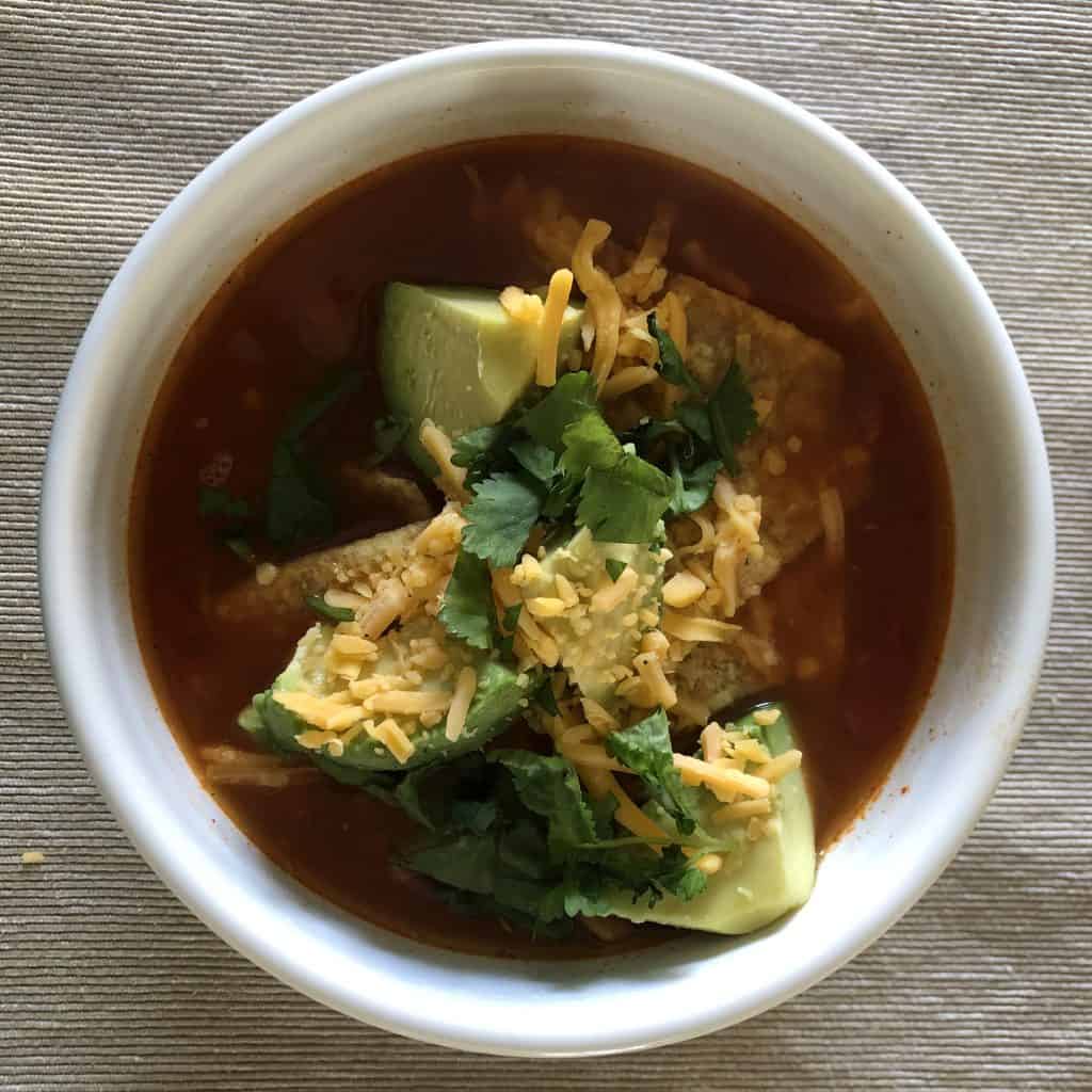 A white bowl of chicken tortilla soup with avocado, shredded cheese, tortilla chips, and cilantro.