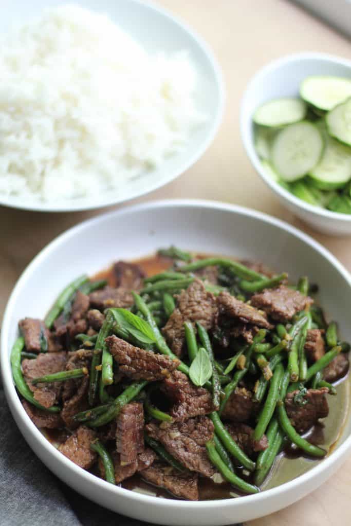 A bowl with beef and green beans in the foreground with bowls of cucumber slices and rice in the background.