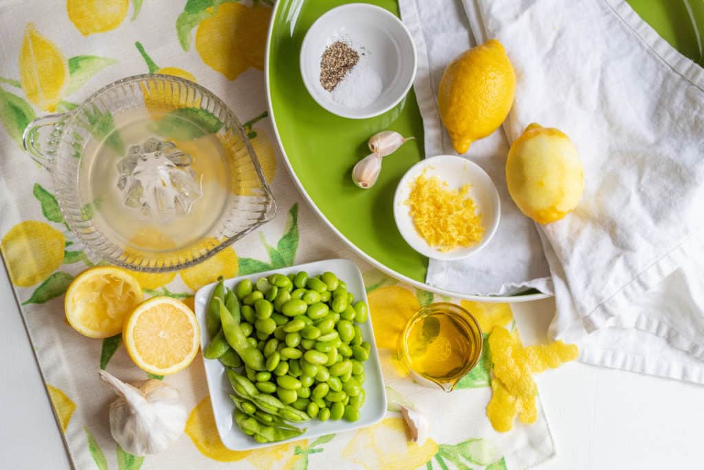 A table with ingredients for an edamame and lemon spread over a round green platter and a lemon placemat. 