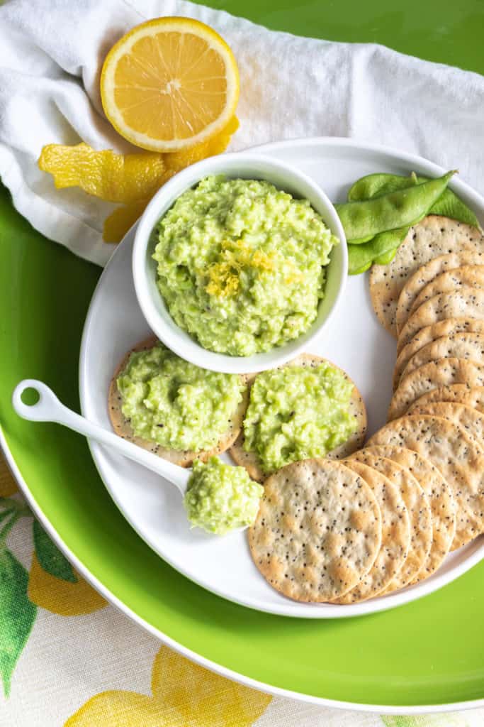 A round plate with crackers and edamame lemon spread with a lemon half in the top left corner.