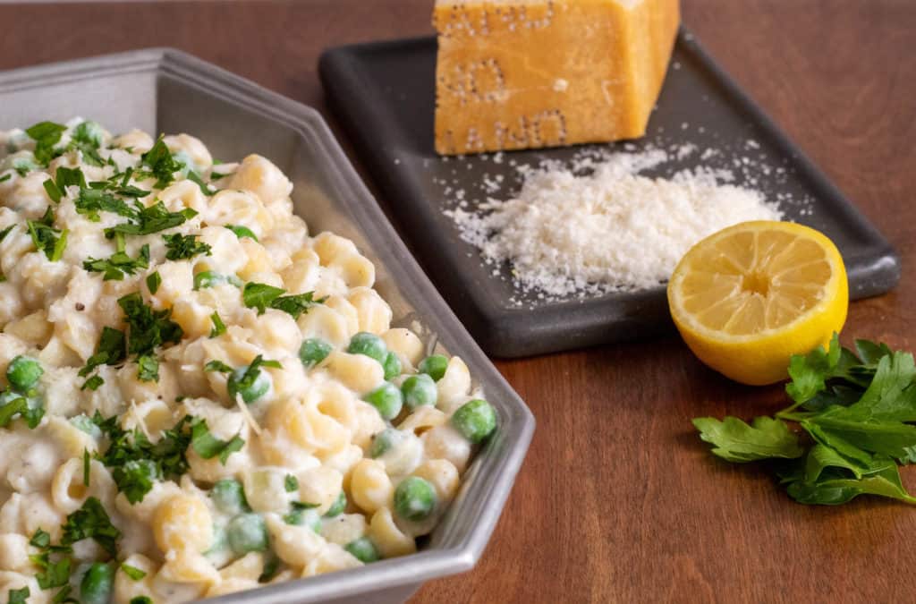 A silver tray of lemon ricotta shell pasta with peas alongside a dish of Parmesan cheese, a half lemon, and fresh parsley.