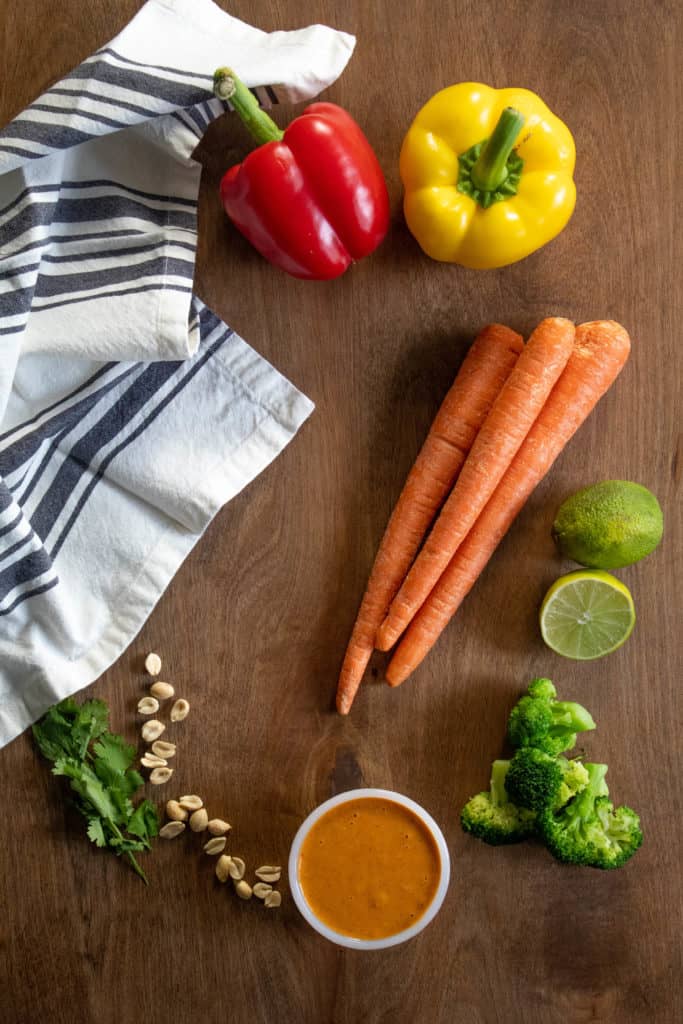 An arrangement of ingredients for Sheet Pan Peanut Chicken: peppers, carrots, lime, broccoli, peanuts, cilantro, and peanut sauce.