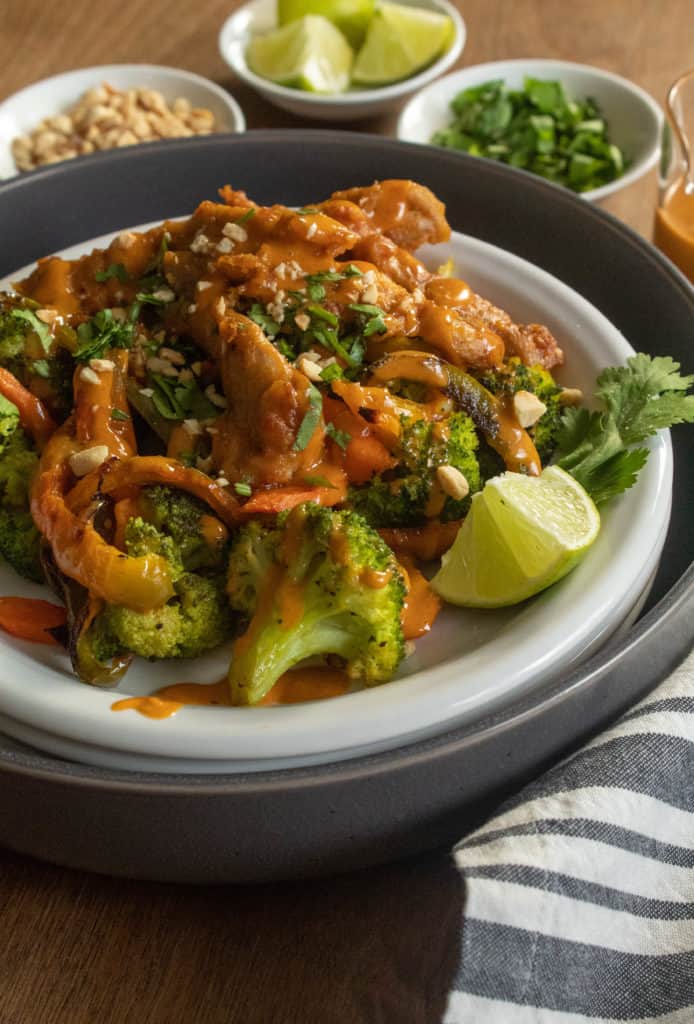 A plate of chicken and vegetables with peanuts, cilantro, and peanut sauce on top.  Small bowls of toppings are in teh background.