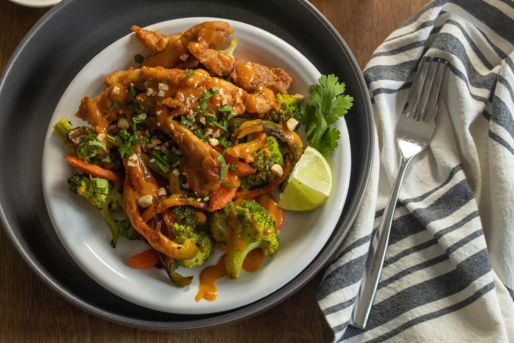 A white bowl of chicken and vegetables alongside a napkin and fork.