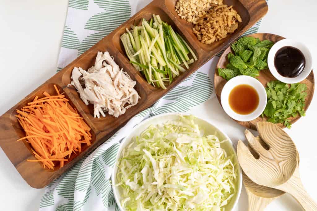 A wooden tray with vegetables and chicken sits next to a bowl of cabbage and a plate of herbs and sauces.