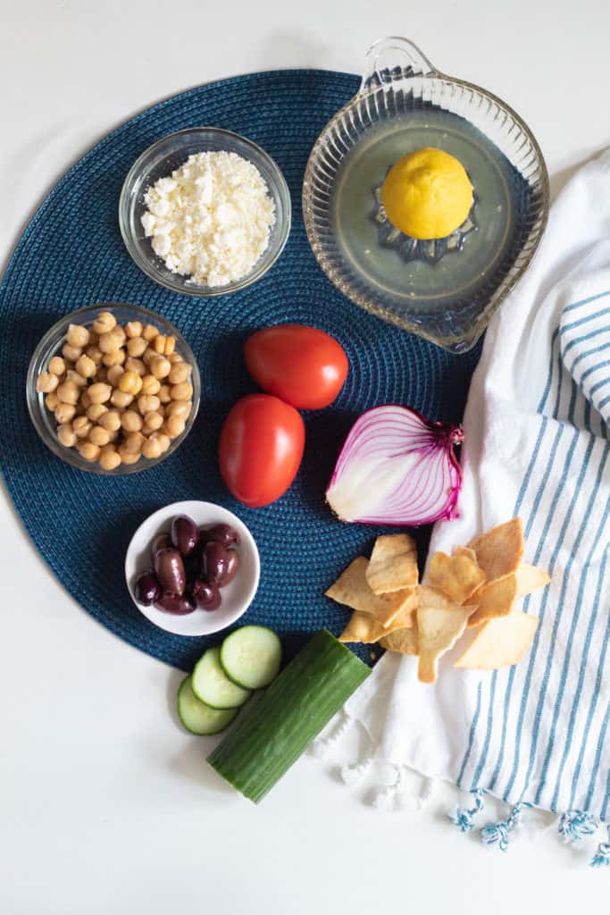 The ingredients for Greek Salad Dip arranged on a blue circle with a blue and white towel.