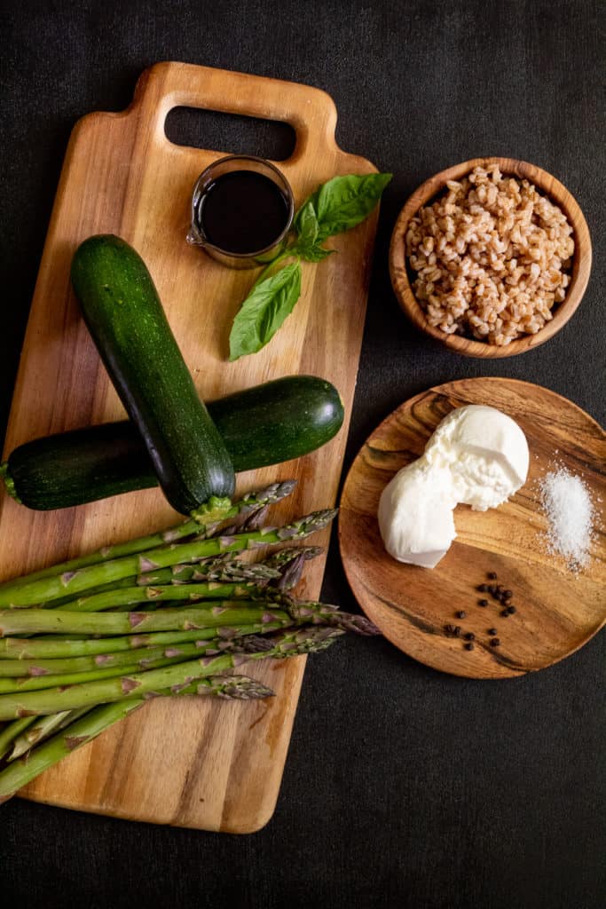 Ingredients, including asparagus, zucchini, basil, balsamic vinegar, burrata, farro, and salt and pepper arranged in various wooden plates over a black background.