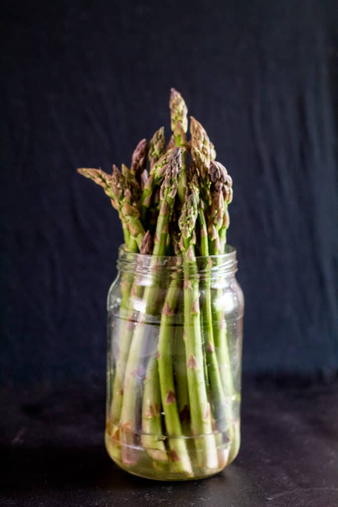 A jar holding a bunch of asparagus upright with a black background.