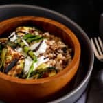 A close-up shot of a brown wooden bowl with farro, zucchini, asparagus, and burrata cheese drizzled with balsamic glaze.