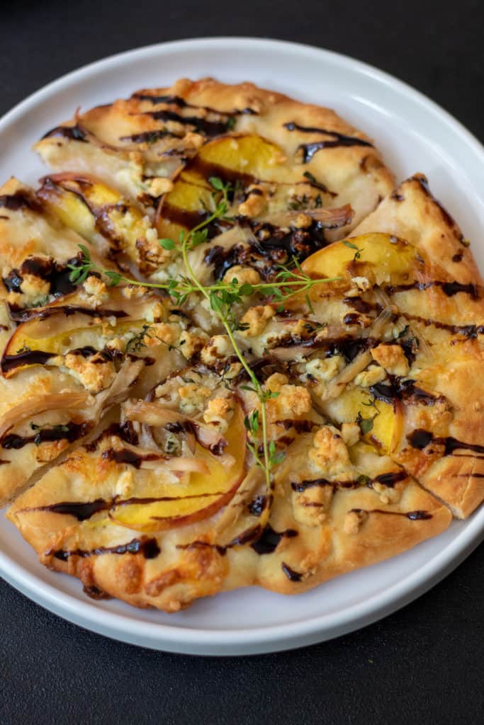 A white plate on a black background. The plate has a nectarine and blue cheese pizza with thyme on it and is drizzled with balsamic vinegar.