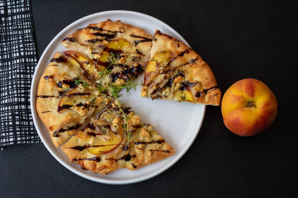 A white plate on a black background. The plate has a nectarine and blue cheese pizza with thyme on it and is drizzled with balsamic vinegar. A nectarine sits alongside the plate.