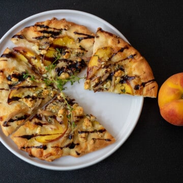 A white plate on a black background. The plate has a nectarine and blue cheese pizza with thyme on it and is drizzled with balsamic vinegar. A nectarine sits alongside the plate.