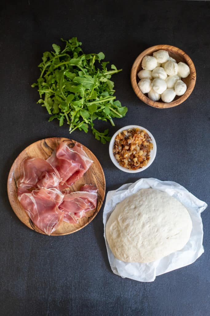 Ingredients for Caramelized Onion and Prosciutto Pizza are arranged on a black surface.