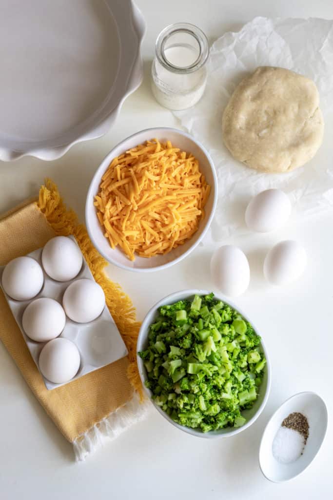An array of ingredients for the preparation of broccoli and cheddar quiche on a white surface.
