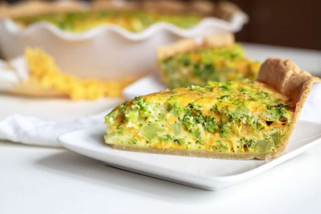 A close-up photo of a slice of broccoli and cheddar quiche on a white square plate.