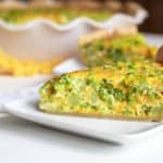 A close-up photo of a slice of broccoli and cheddar quiche on a white square plate.