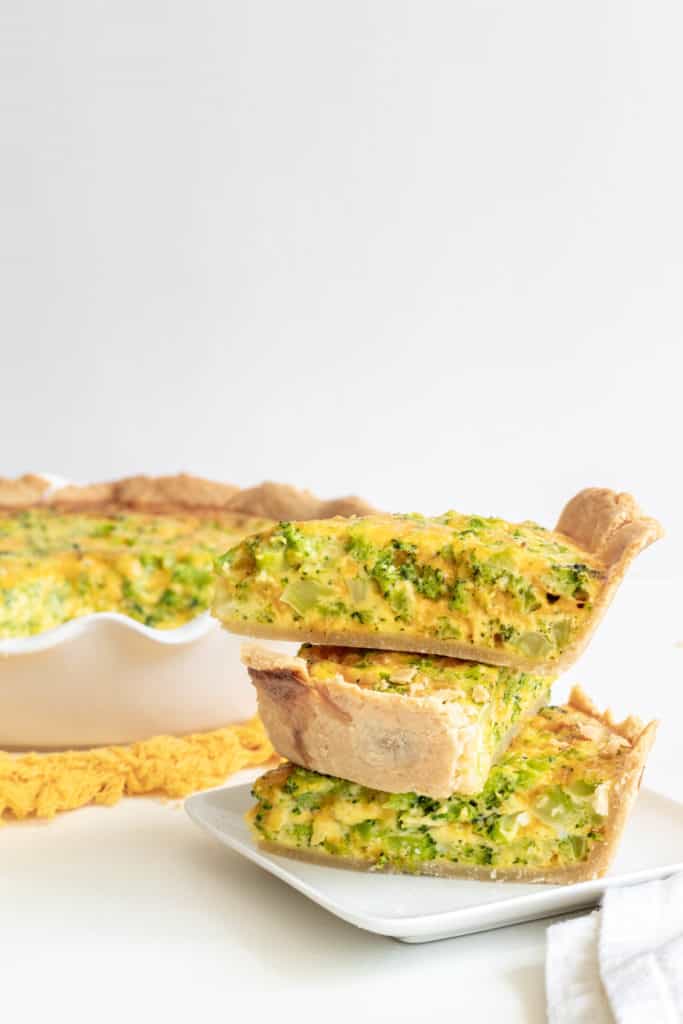 A stach of quiche slices on a white plate with a baked quiche in the background.