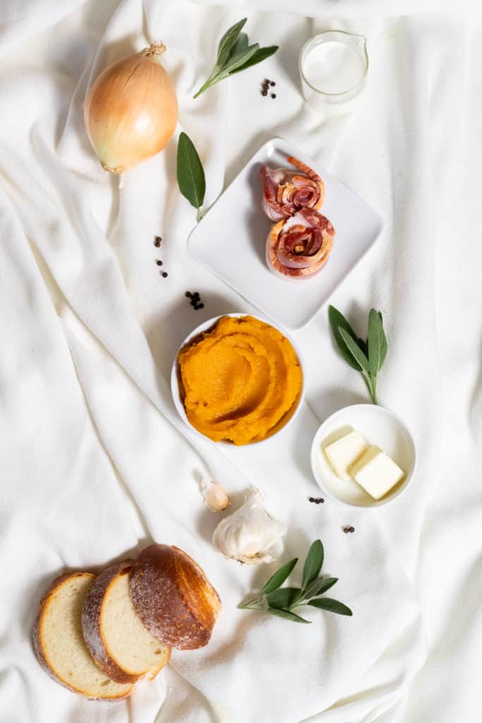 Ingredients for Pumpkin and Bacon Bisque are arranged on a white fabric surface.