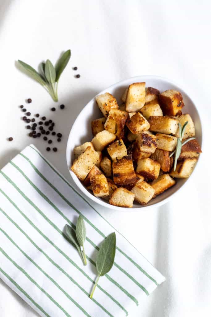 A white bowl of bacon- and sage-infused croutons on a white surface with sage and peppercorns scattered around.