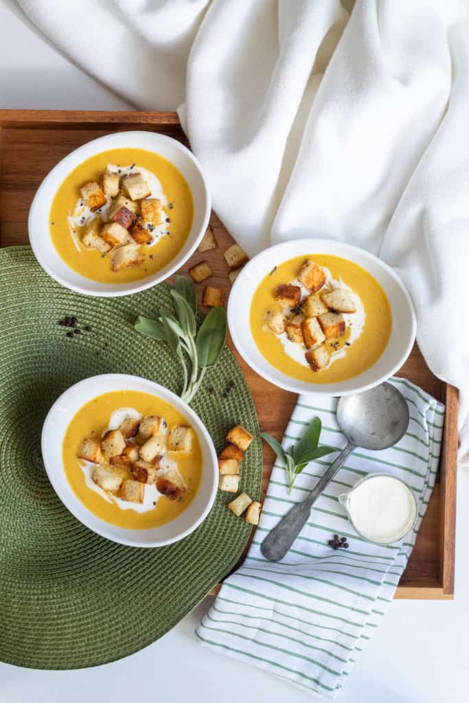 Three white bowls of Pumpkin and Bacon Bisque are on a wooden tray with sage and croutons scattered around.