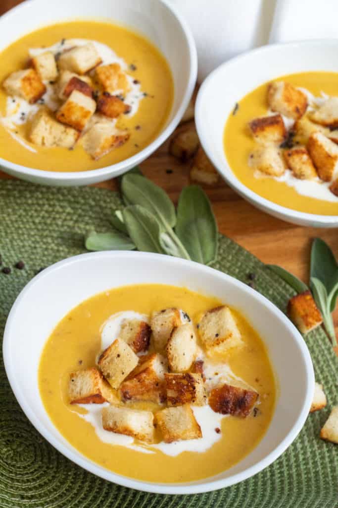 Three white bowls of Pumpkin and Bacon Bisque are on a wooden tray with sage and croutons scattered around.