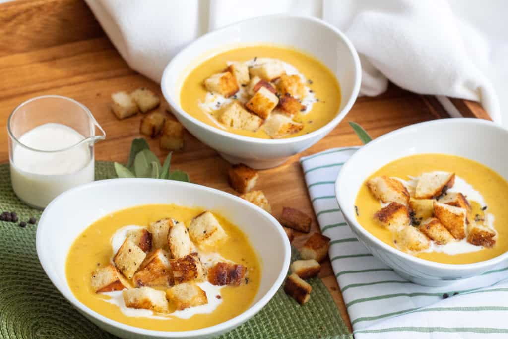 Three white bowls of yellow soup with croutons are on a wooden tray with sage and croutons scattered around.