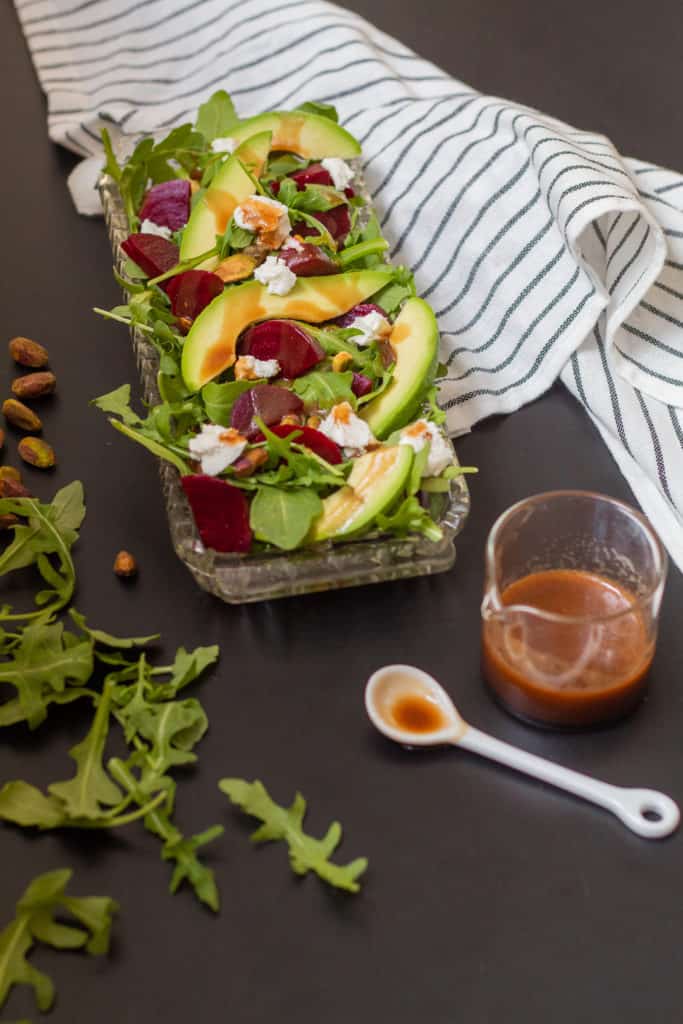 A beet and avocado salad in a glass dish sits on a black surface. A spoonful of chili-soy vinaigrette is nearby next to a striped black and white towel.