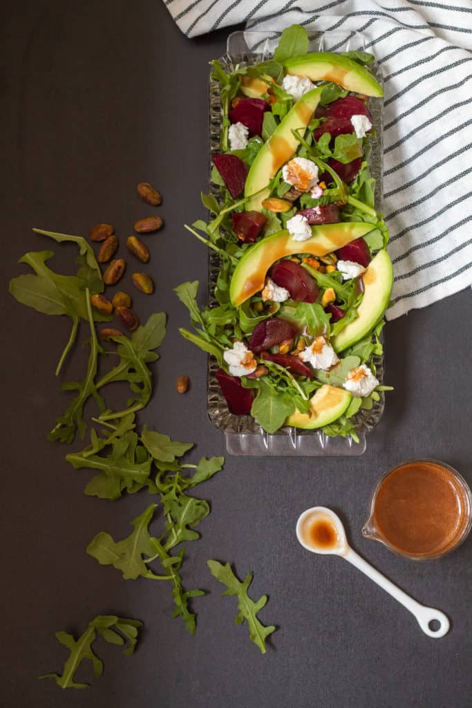 A beet and avocado salad in a glass dish sits on a black surface. A spoonful of chili-soy vinaigrette is nearby next to a striped black and white towel.