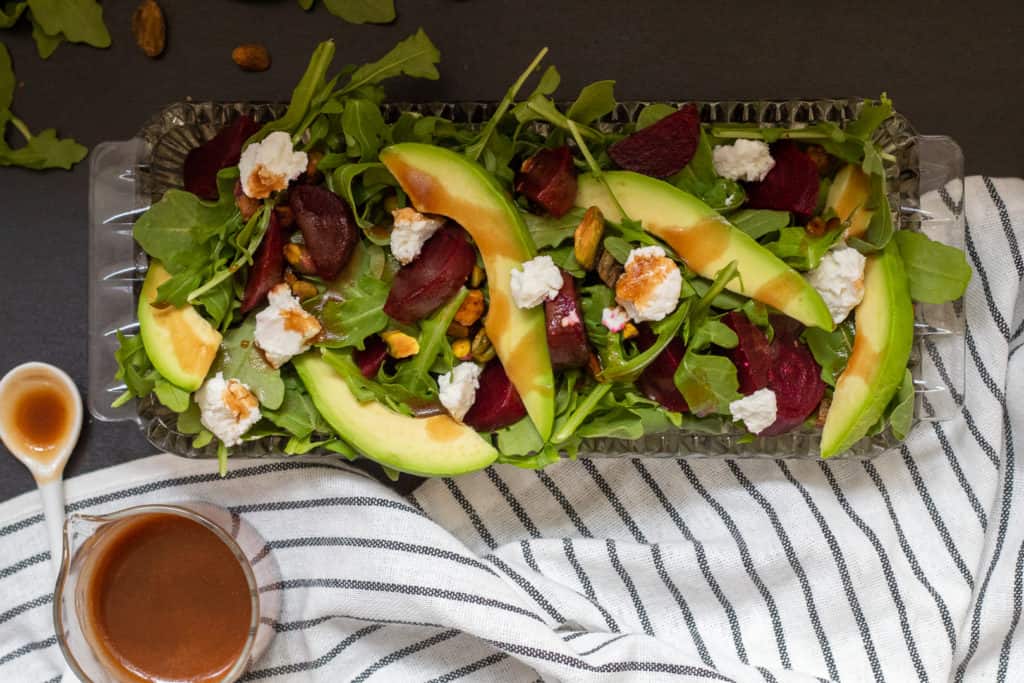 A beet and avocado salad in a glass dish sits on a black surface. A spoonful of chili-soy vinaigrette is nearby next to a striped black and white towel.