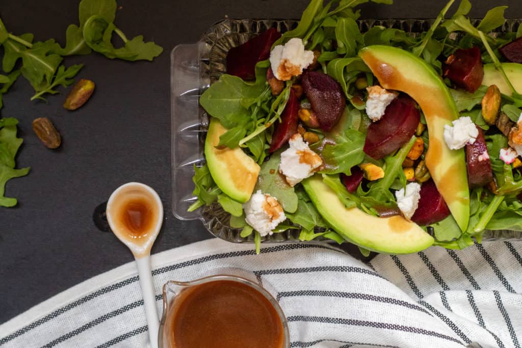 A beet and avocado salad in a glass dish sits on a black surface. A spoonful of dressing is nearby next to a striped black and white towel.