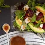 A beet and avocado salad in a glass dish sits on a black surface. A spoonful of dressing is nearby next to a striped black and white towel.