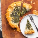 A Spinach and Feta Pie sits on a wooden cutting board with a serving on a white plate next to it.