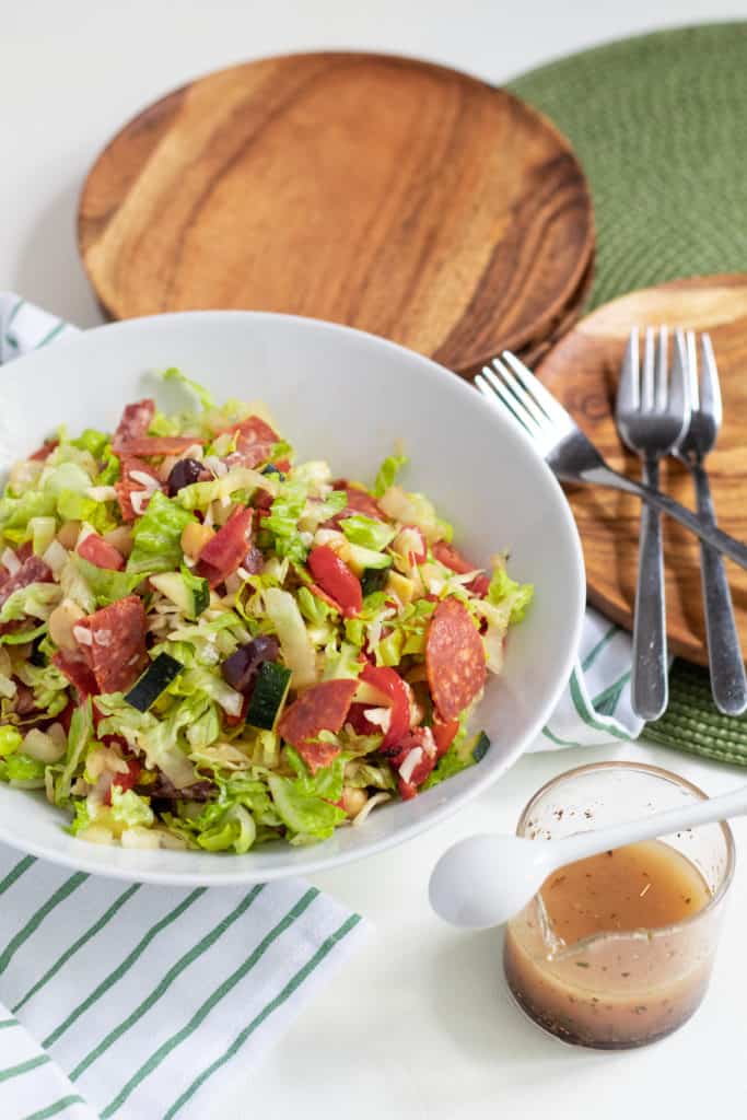 A chopped salad in a white bowl is surrounded by wooden plates, a jar of salad dressing, and forks on a white and green background.