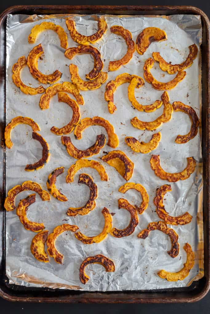 Half-moon pieces of roasted squash are scattered on a foil-lined baking tray.