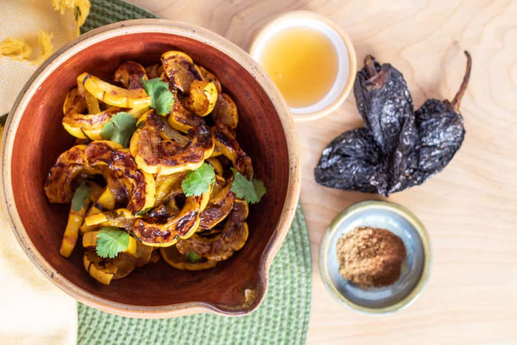 A bowl of golden squash is on teh left of the image. Small bowls of honey and spices are to the right of the bowl, next to two dried chiles.