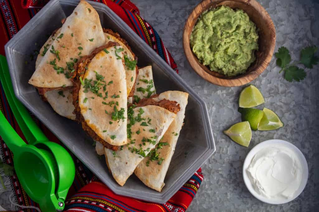 Baked Sweet Potato and Soyrizo Tacos are arranged in a silver serving dish. Guacamole, limes, and sour cream are arranged on a silver surface nearby.