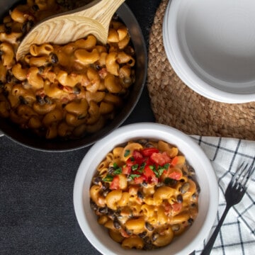 A pot of Vegan Chili Mac sits on a black surface next to a stack of white bowls and a serving of pasta garnished with chopped fresh tomatoes.