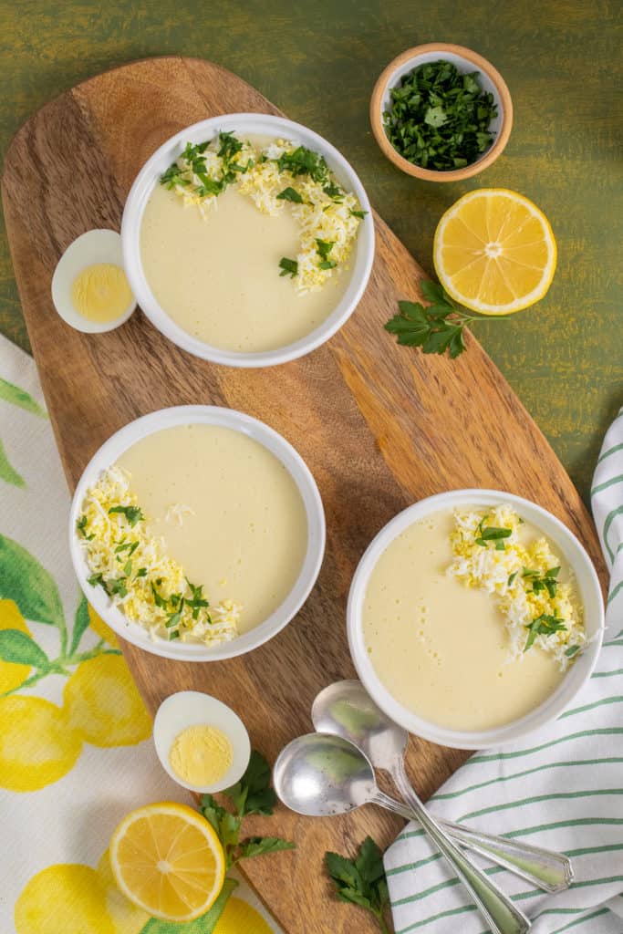 Three bowls of pale yellow soup are arranged on a wooden platter with lemons, hard boiled eggs, and parsley alongside.