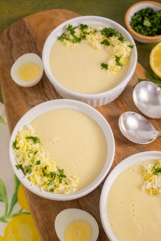 Bowls of Avgolemono Soup are garnished with grated hard boiled egg and parsley. They sit on a wooden tray over a green surface.