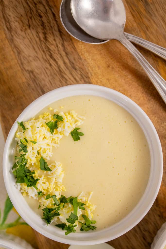 A white bowl of pale yellow soup sits on a wooden background with two silver spoons alongside.