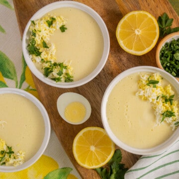 Three bowls of Avgolemono Soup are arranged on a wooden board and over fabric with lemons and leaves.