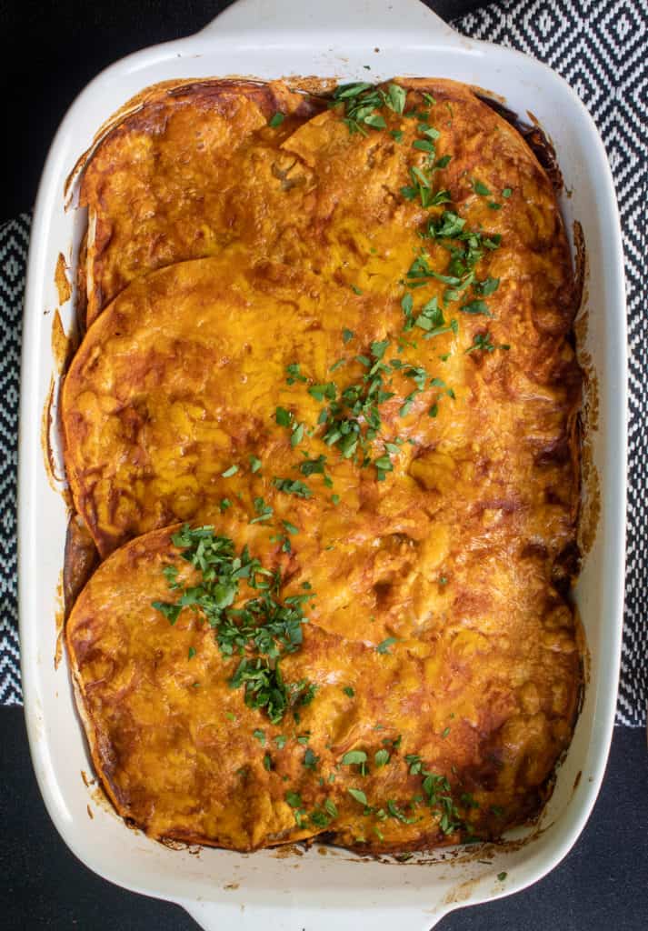 The finished casserole dish of Black Bean and Sweet Potato Stacked Enchiladas sits on a black and white textile background.