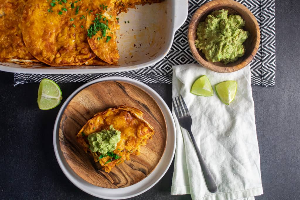 A serving of enchilada on a wood plate sits alongside the baking dish. Garnishes like lime wedges and guacamole are next to the baking dish.