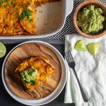 A serving of enchilada on a wood plate sits alongside the baking dish. Garnishes like lime wedges and guacamole are next to the baking dish.