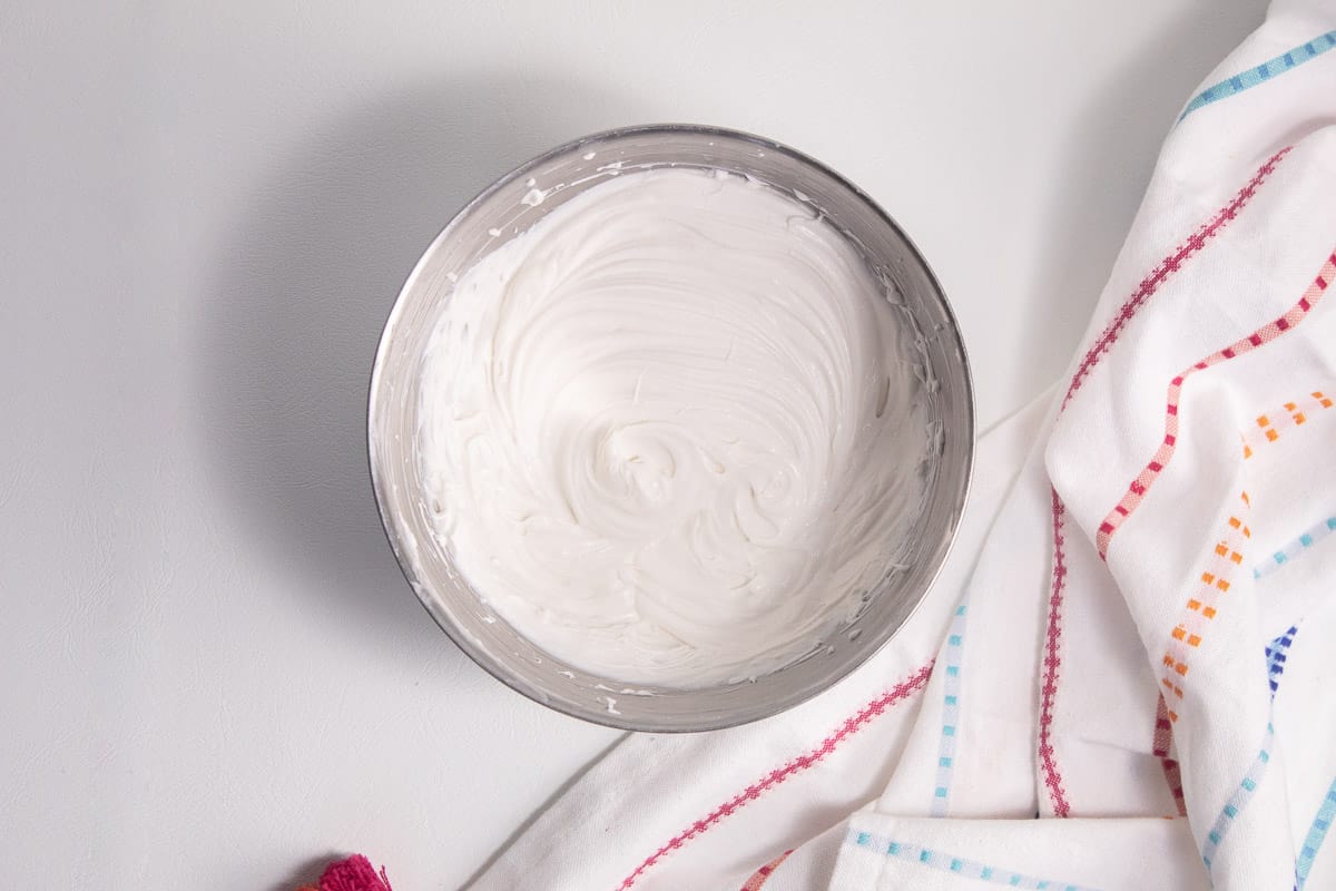 Cream cheese and coconut milk are whipped to a smooth consistency in a stainless steel bowl over a white surface.