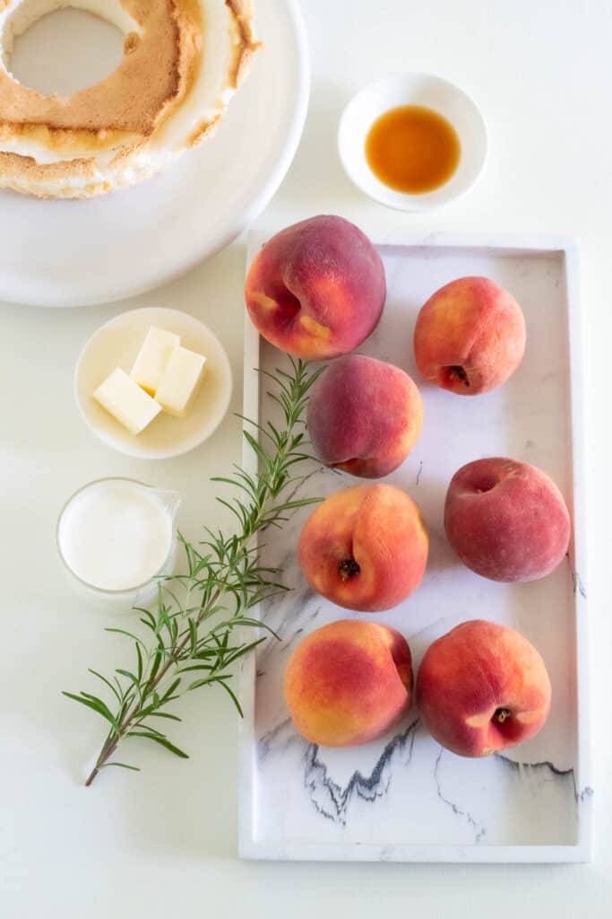Ingredients for Fresh Peach Trifle with Angel Food Croutons and Rosemary Syrup: Peaches, a rosemary sprig, cream, butter, vanilla, and angel food cake are arranged on a white and gray surface.