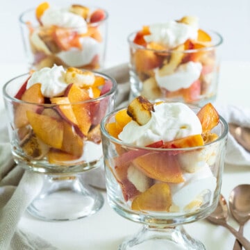 Four glasses filled with sliced fresh peaches, whipped cream, and angel food croutons arranged on a white surface.