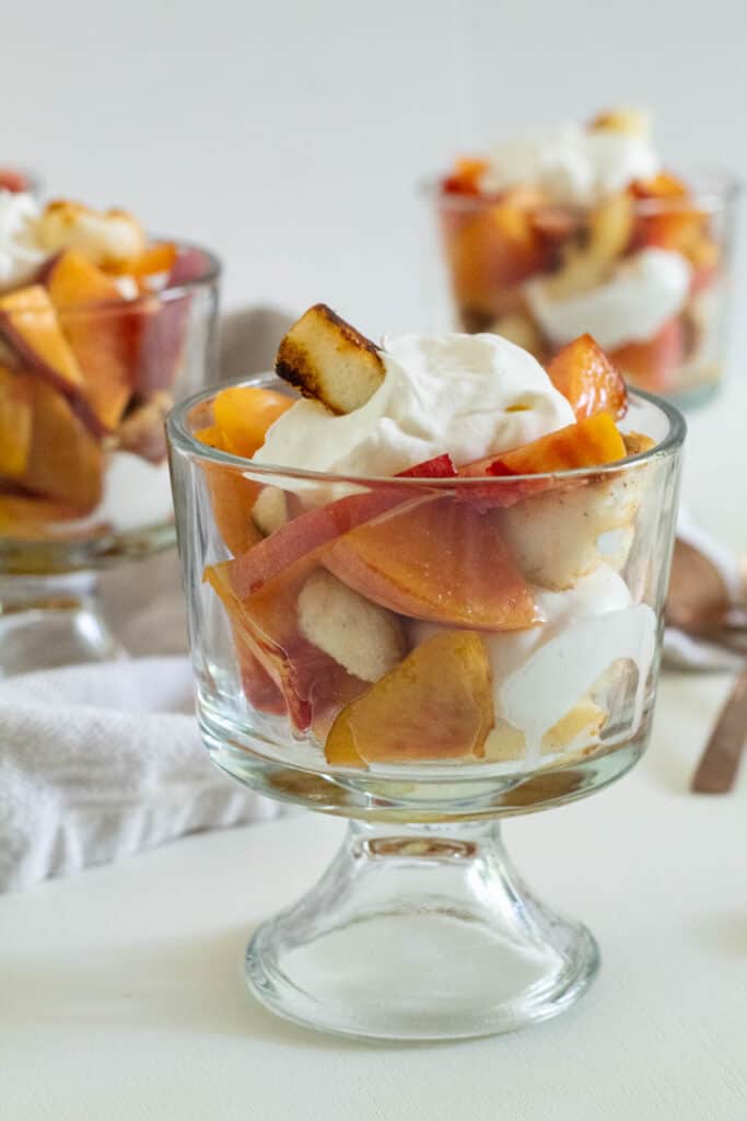 A serving of sliced peaches, whipped cream, and angel food croutons on a white surface with additional servings in the background.