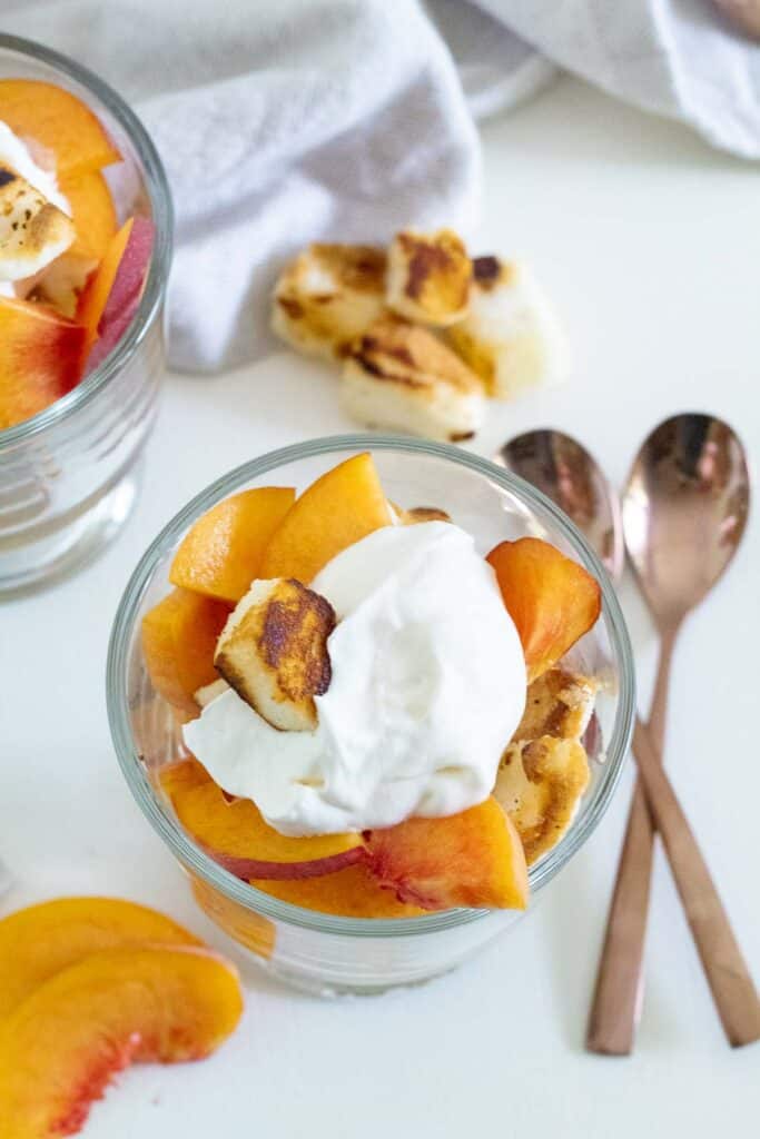 A serving of sliced peaches, whipped cream, and angel food croutons on a white surface with two spoons and sliced peached in the background.